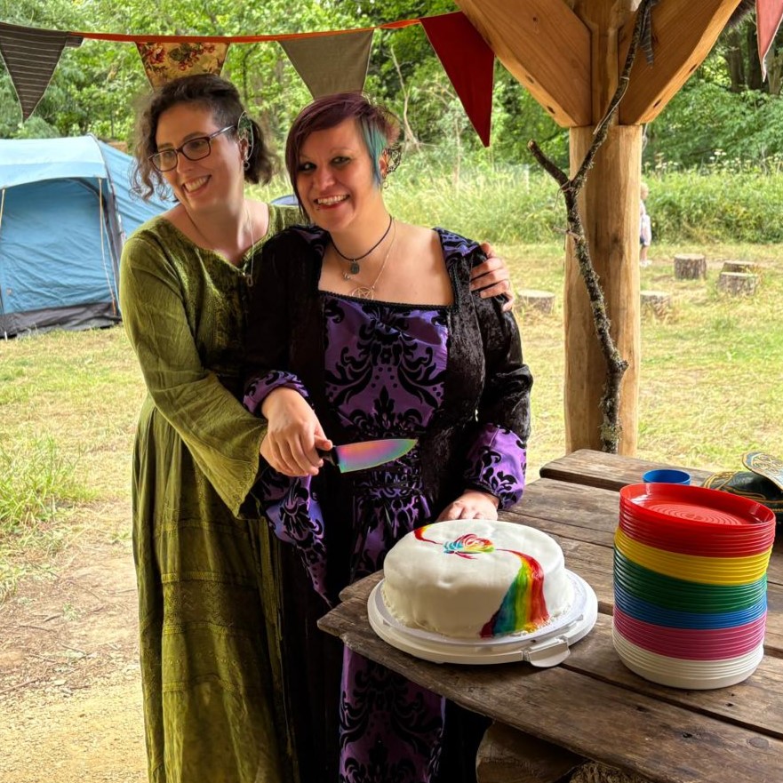 Charlie and I are standing at the wooden roundhouse at Tortworth Arboretum, cutting a large white wedding cake with a rainbow rose on the wedding cake with a rainbow knife. You can see tents in the background behind us. We're both wearing Medieval fantasy style gowns with long skirts and flowing sleeves. Charlie's is purple and black, mine's green