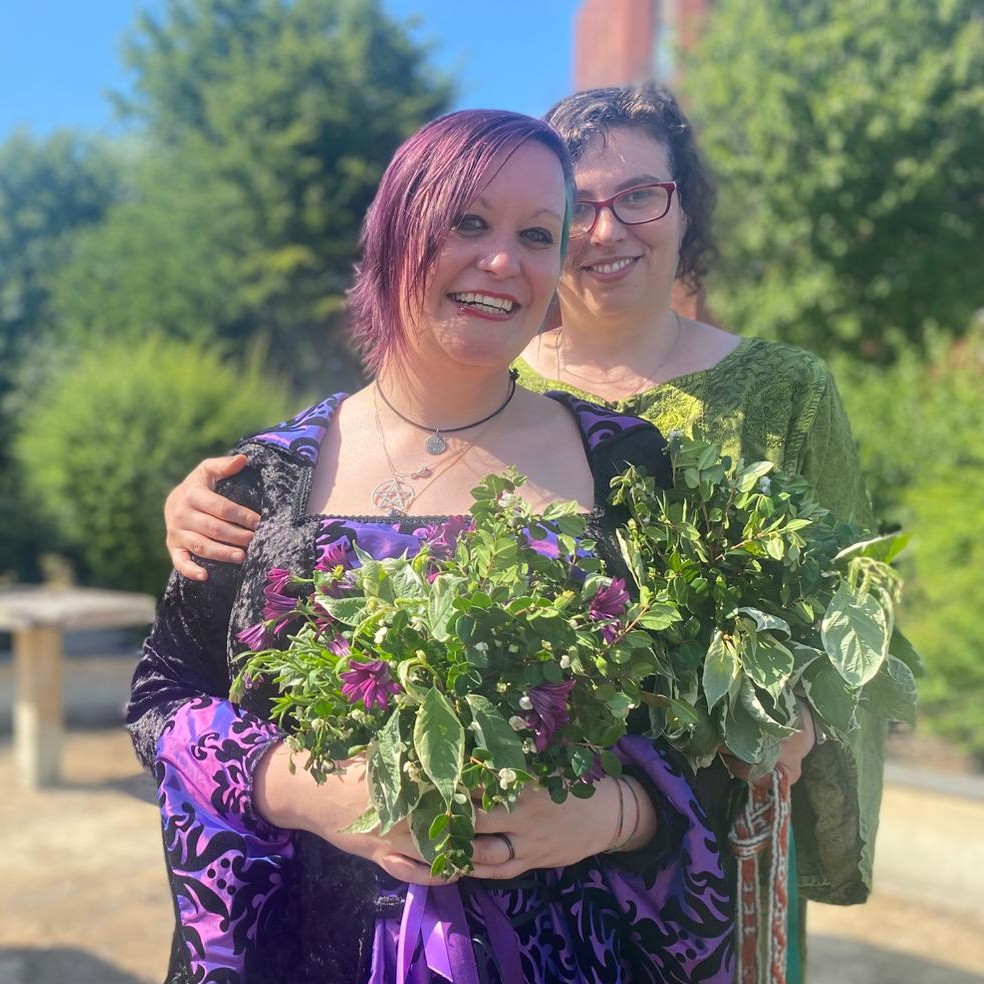 Two women stand beside each other surrounded by verdant foliage under a clear blue sky. Charlie stands in front, she is wearing a long mediaeval style dress of black velvet with a square neckline and a panel of purple brocade down the front which has at least two different tones in it in the sunshine. She has draping teardrop sleeves which show the same purple brocade on the cuffs. She has a pentagram necklace. Purple hair cascades down the right side of her face, and teal hair tucks behind her left ear. She has dramatic eye makeup and her smile is radiant.
I'm standing slightly behind her with my hand on her shoulder. My dress is vivid green with intricate embroidery on the bodice and draping sleeves too. I'm also smiling delightedly. My hair is dark and has gone incredibly frizzy with the heat and humidity that comes from getting married in the middle of an apocalyptic heatwave, but I' m the happiest I've ever been in my life and I don't care.
We're holding bouquets that were grown by my Mum, of purple asters, myrtle flowers and variegated foliage.