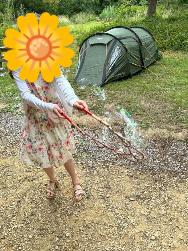 A young girl in a floral dress waves a bubble wand of two sticks joined by a string with multiple loops to make a cascade of bubbles