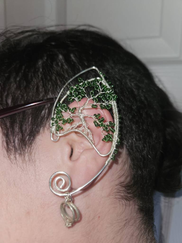 A wirework ear cuff - silver wire that sits on an ear in the shape of a pointed elf ear. Hanging from the bottom is a charm of two wedding rings. The top is covered in a tree wirework design - thin silver wire is twisted together like roots turning into a tree trunk, then splits apart to make the branches. Green glass beads form the leaves