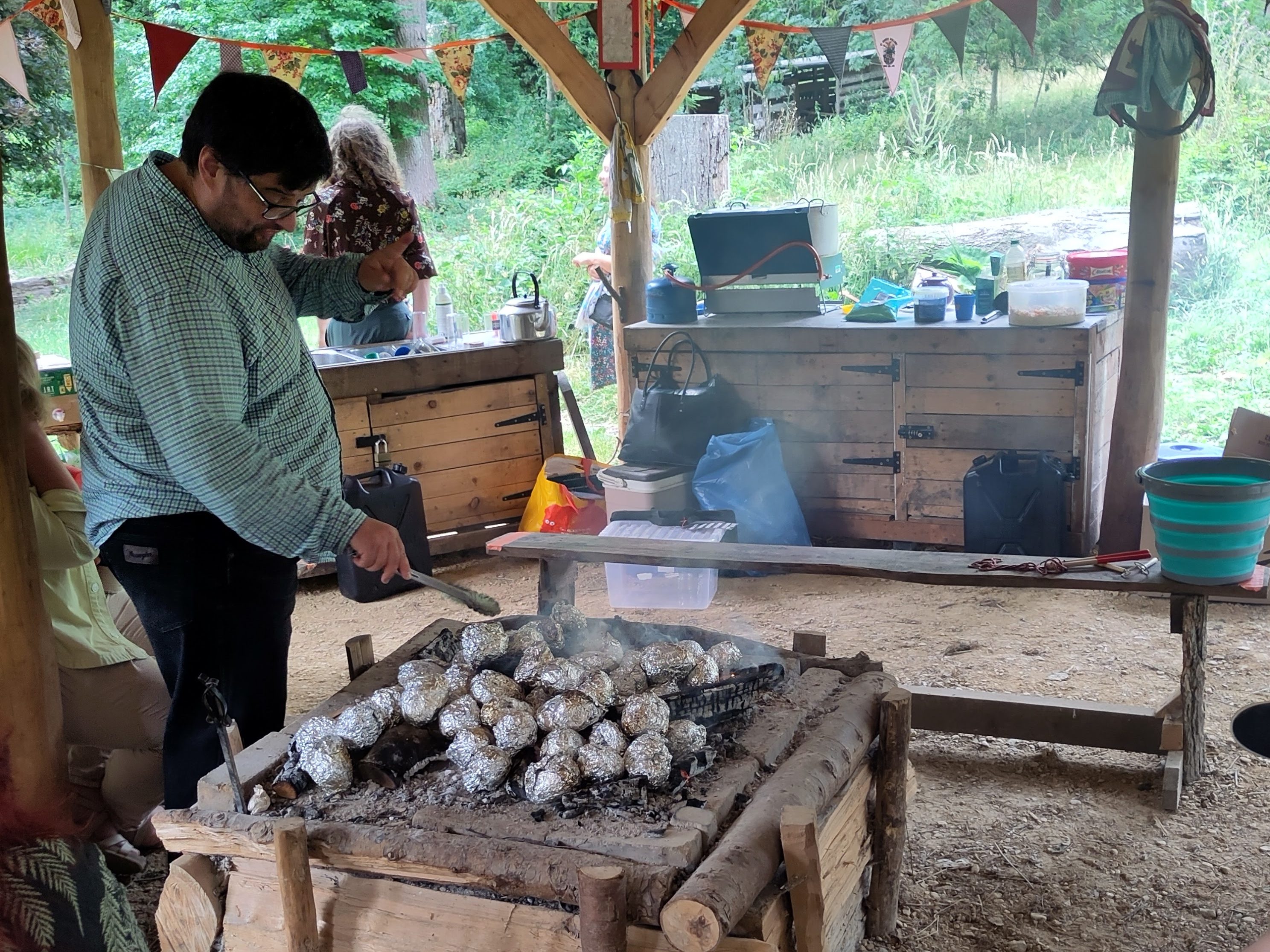 A friend of ours, a dark haired, bearded man with a green shirt, rotates the foil wrapped jacket potatoes with tongs on embers in the fire pit
