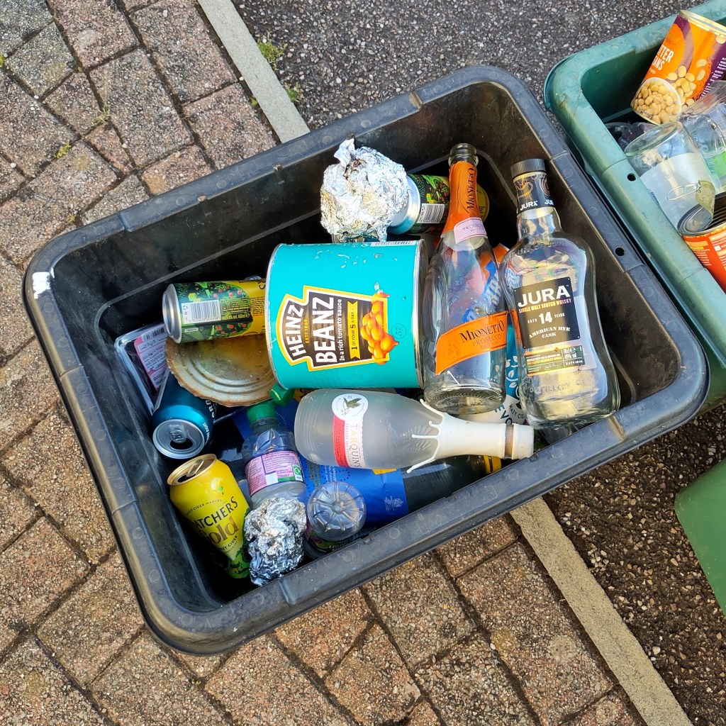 A kerbside recycling bin containing a caterings sized tin of baked beans, balls of slightly charred aluminium foil, cans of cider and bottles of whisky, gin and prosecco.