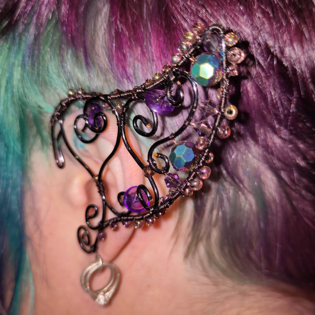 A wirework ear cuff in curls of black wire, in the shape of a pointed elf ear and with a wedding ring charm hanging off the bottom. It is decorated with iridescent beads in purple and teal, matching the purple teal of Charlie's hair.