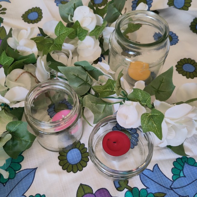 Three glass jars on a vintage tablecloth with different coloured tealights in them and sprays of plastic wisteria round them