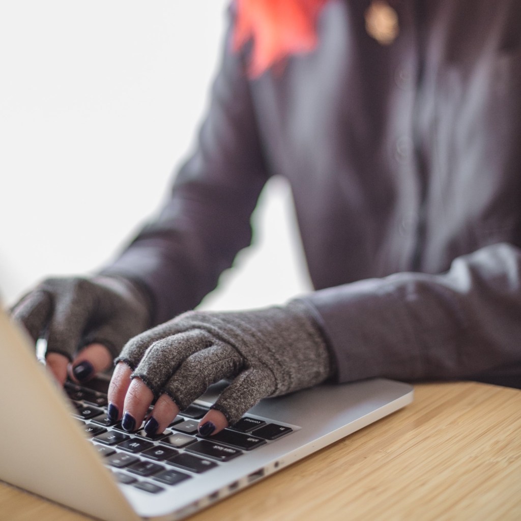 A disabled Asian genderfluid person types on a laptop while wearing compression gloves. The hands and keyboard are the focal point.