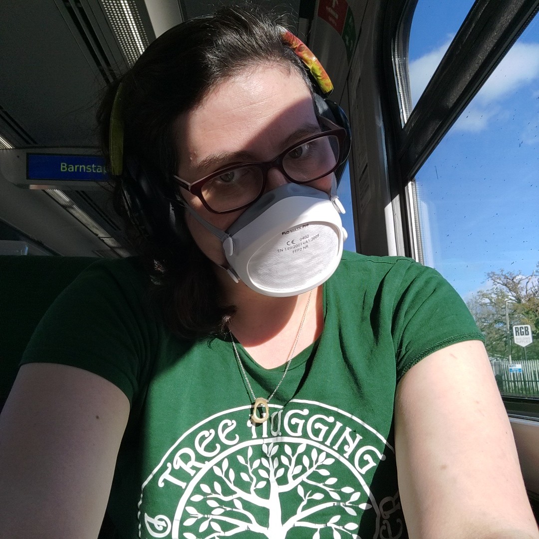 Myself, a white woman with dark hair, glasses, headphones held together with colourful duck tape and a flo mask, sitting on a train