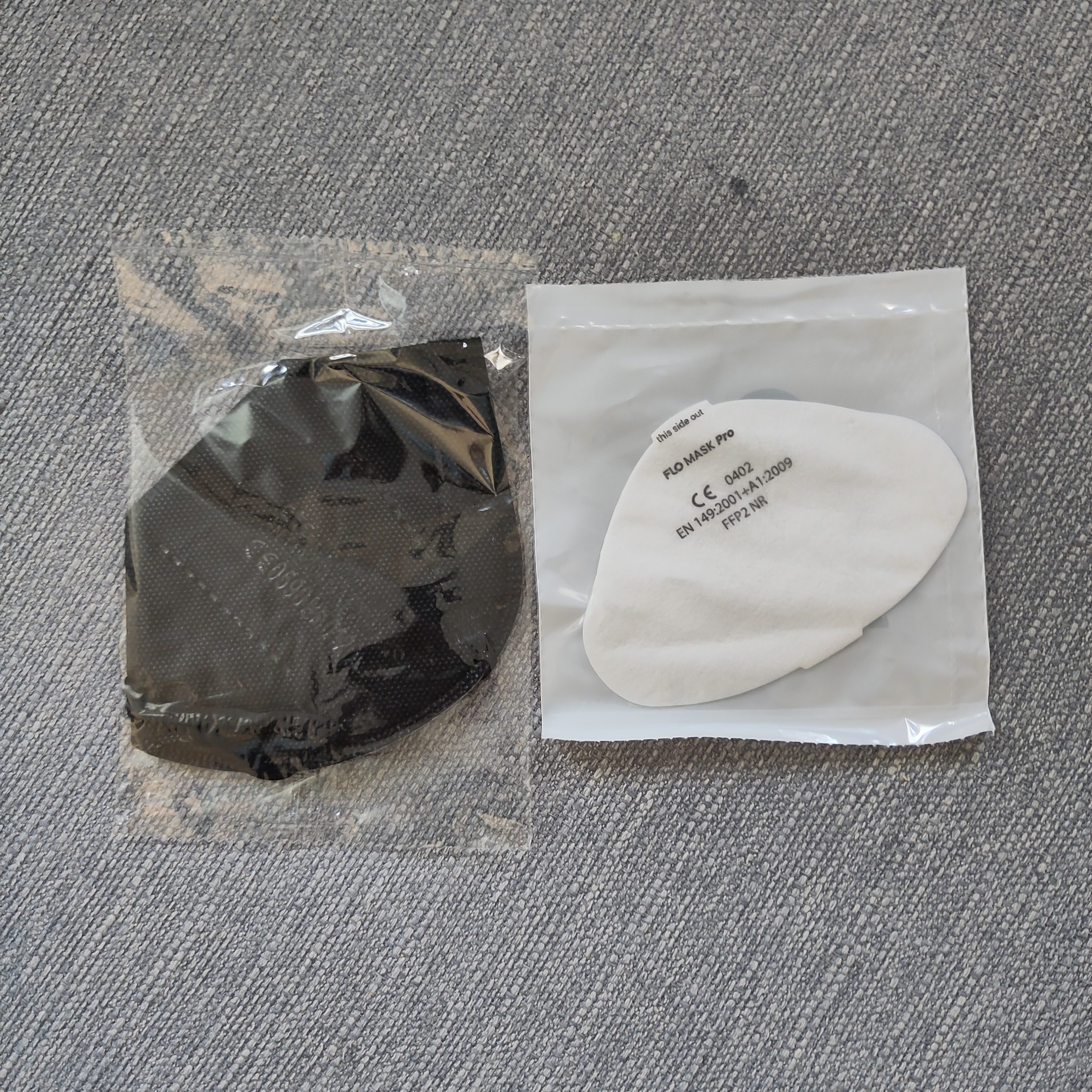 An FFP2 mask and a Flo mask insert side by side, showing that the disposable mask is slightly larger than the insert and made of two layers of plastic and also that the packaging it comes in is larger
