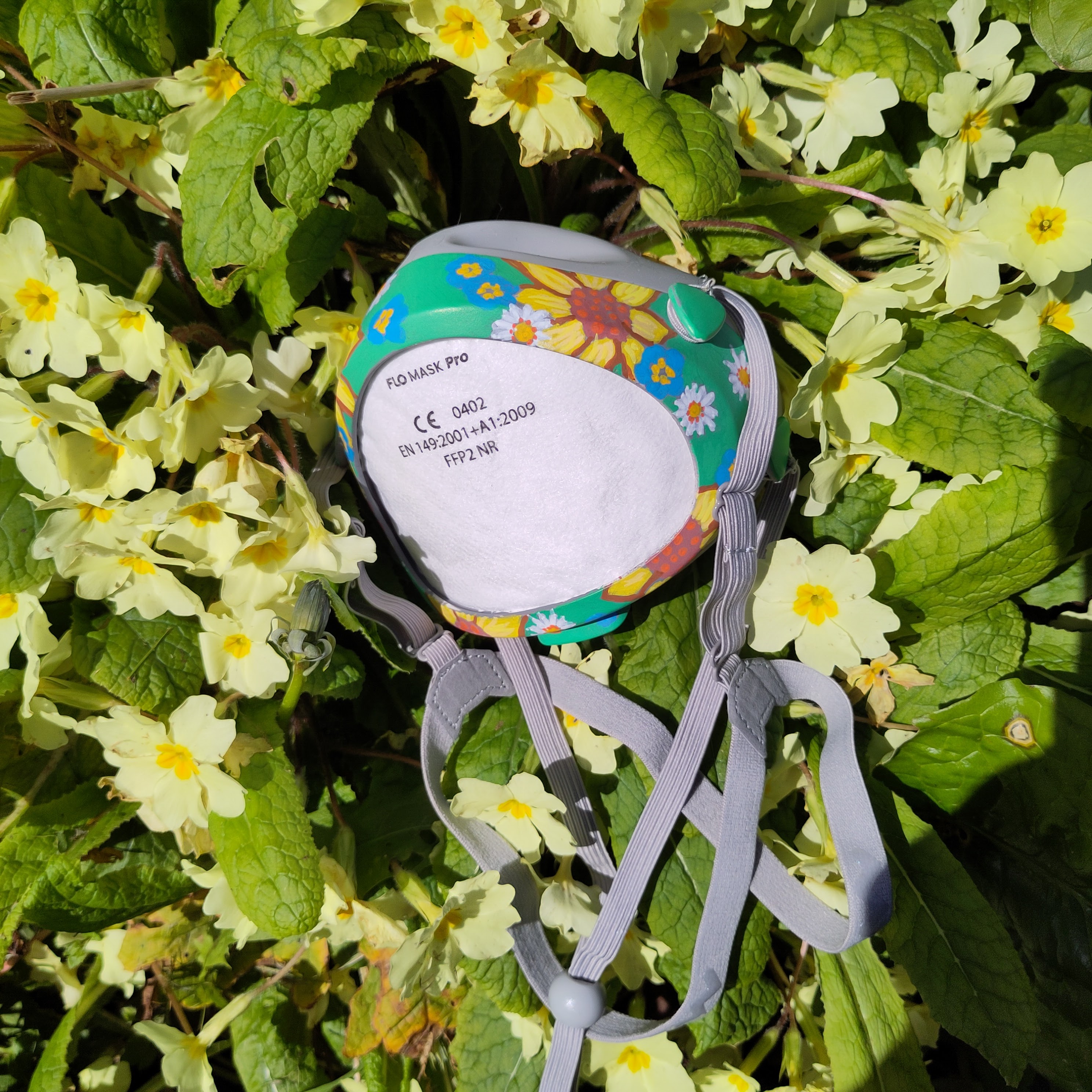 A flo mask with stylised sunflowers, forget me nots and daisies painted on in acrylic paint, lying on a bed of primroses 